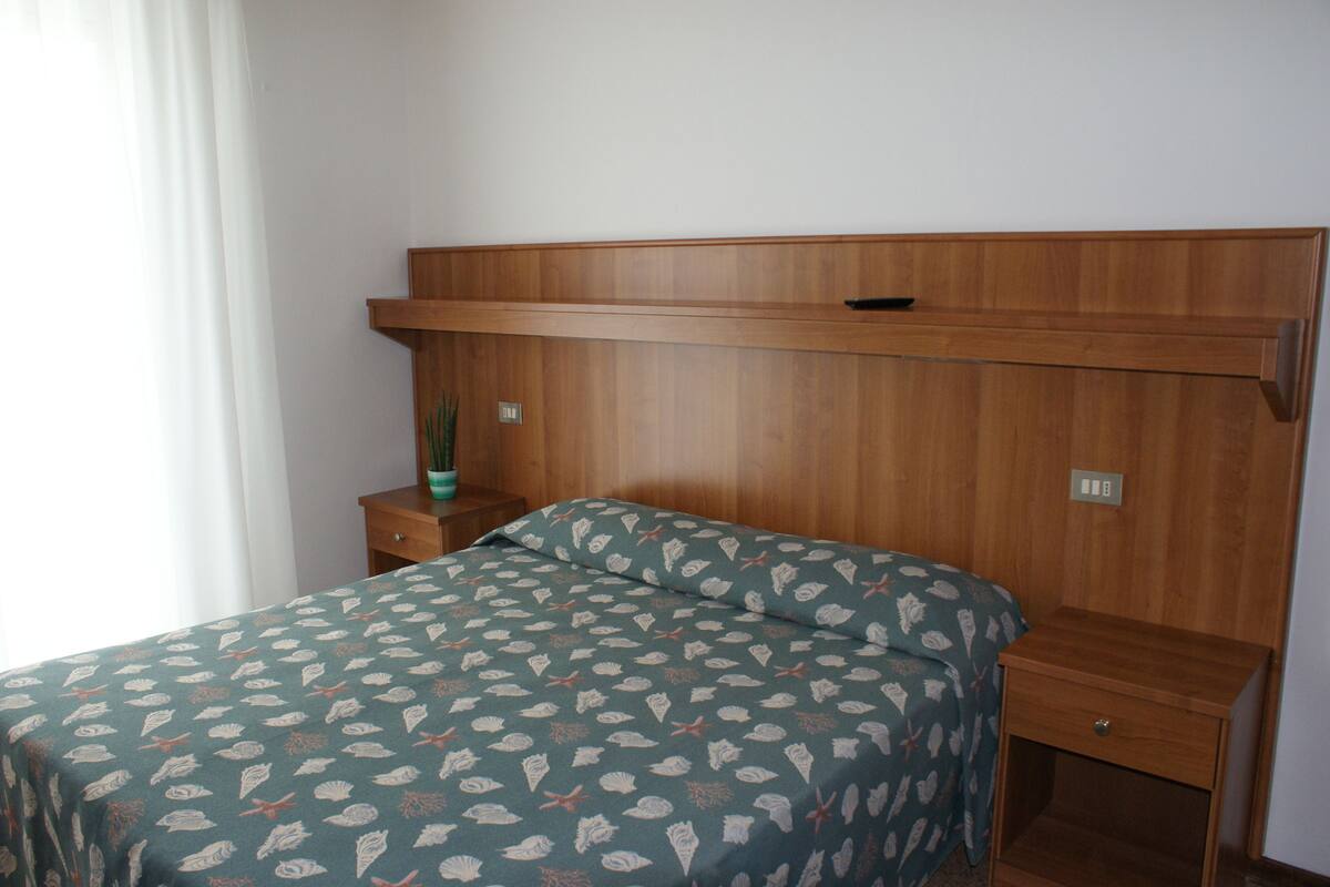 A bedroom with a wooden headboard, a bed with a patterned bedspread, a nightstand with a plant, and a small drawer.
