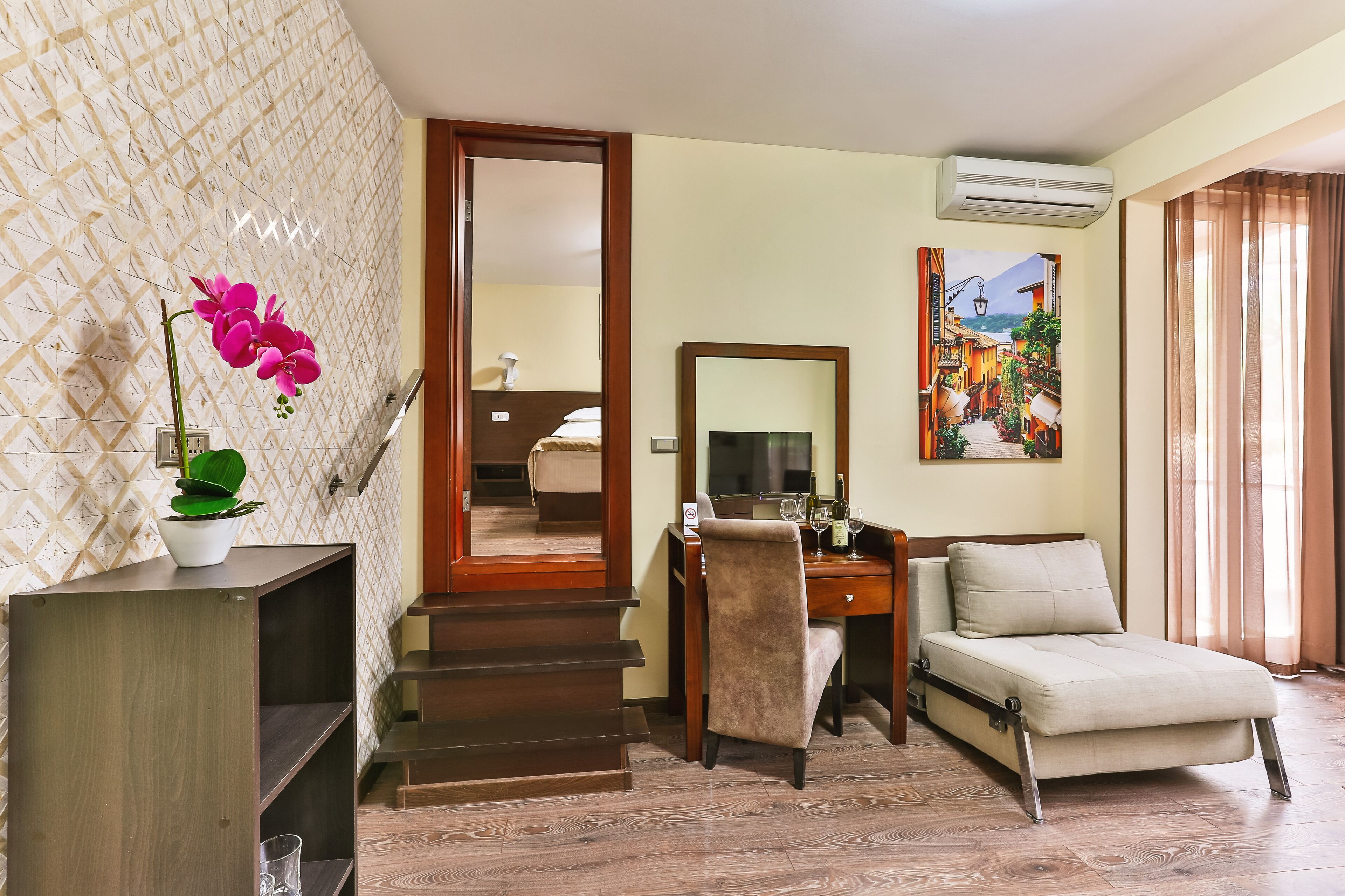 A hotel room with a wooden desk, a sofa, a mirror, a potted plant, and a painting of a street scene.