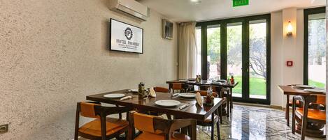 Un comedor de hotel con mesas dispuestas para el servicio, un cartel enmarcado en la pared y vistas al exterior a través de grandes ventanales.