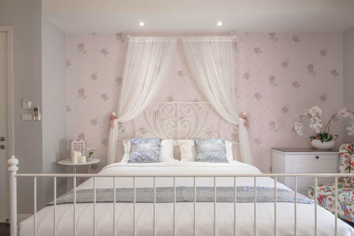 A bedroom with a white bed, floral wallpaper, and a white nightstand with a lamp.