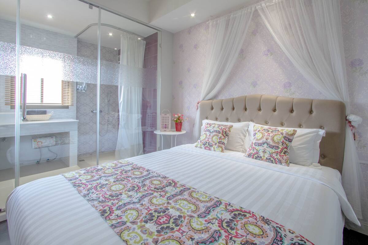 A bedroom with a large bed, a glass shower enclosure, a bedside table with a lamp, and a wall with floral wallpaper.