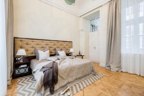 A bedroom with a tufted headboard, a bed with a beige blanket, a wooden nightstand with a lamp, and a patterned rug. - Residence Wollzeile (Vienna)