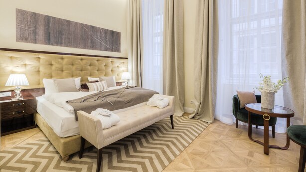 A hotel room with a large bed, a bedside table with a lamp, a chair, a small table with a vase, and a view of buildings through sheer curtains. - Residence Wollzeile (Vienna)