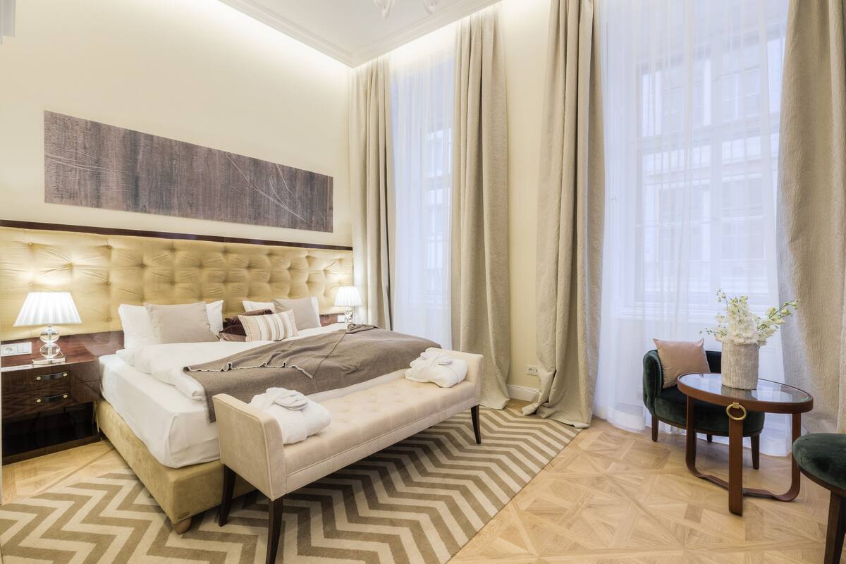 A hotel room with a large bed, a bedside table with a lamp, a chair, a small table with a vase, and a view of buildings through sheer curtains.