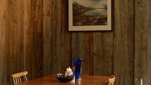Une table à manger en bois, entourée de chaises, un tableau représentant un paysage encadré, un vase bleu et une bougie allumée.