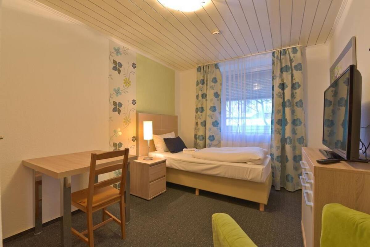A small, well-lit room with a bed, a desk, a chair, and a television.