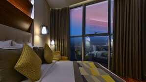 A hotel room with a large bed, a sofa, a lamp, and a view of the city at night. - The Avenue Hotel (Doha)