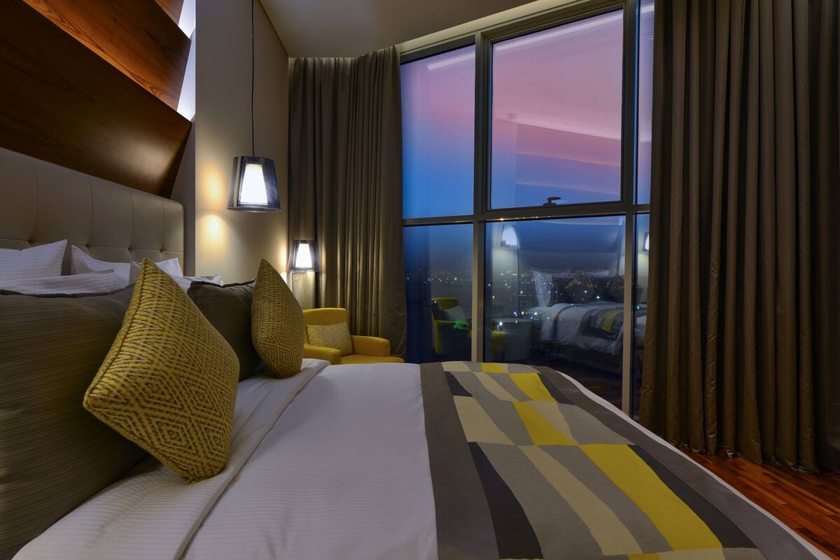 Een hotelkamer met een groot bed, een bank, een lamp en uitzicht op de stad 's nachts.