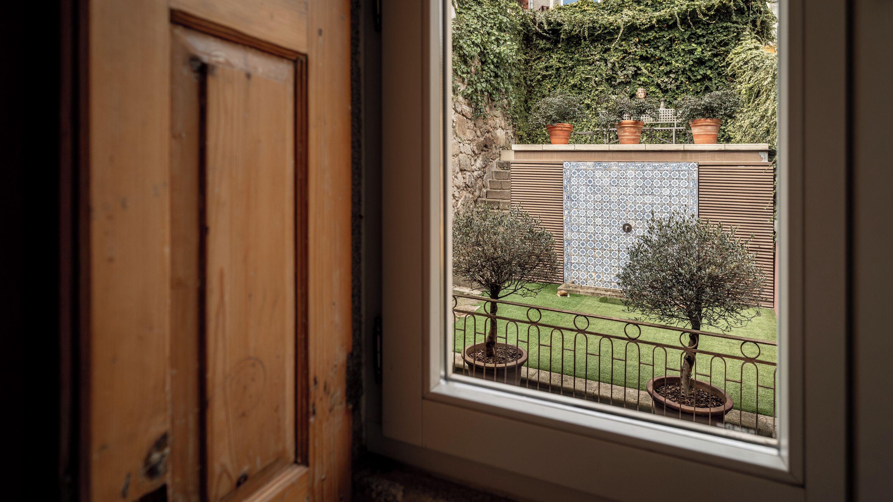 A window view of a garden with potted trees, a stone wall, and a tiled wall.