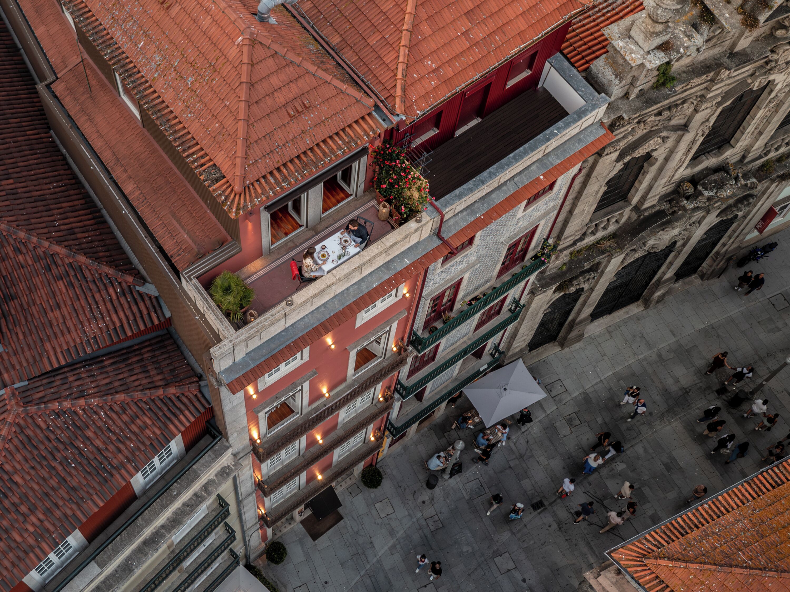 Vista aérea de um edifício histórico com telhado de telhas vermelhas, várias varandas e um pátio com pessoas.