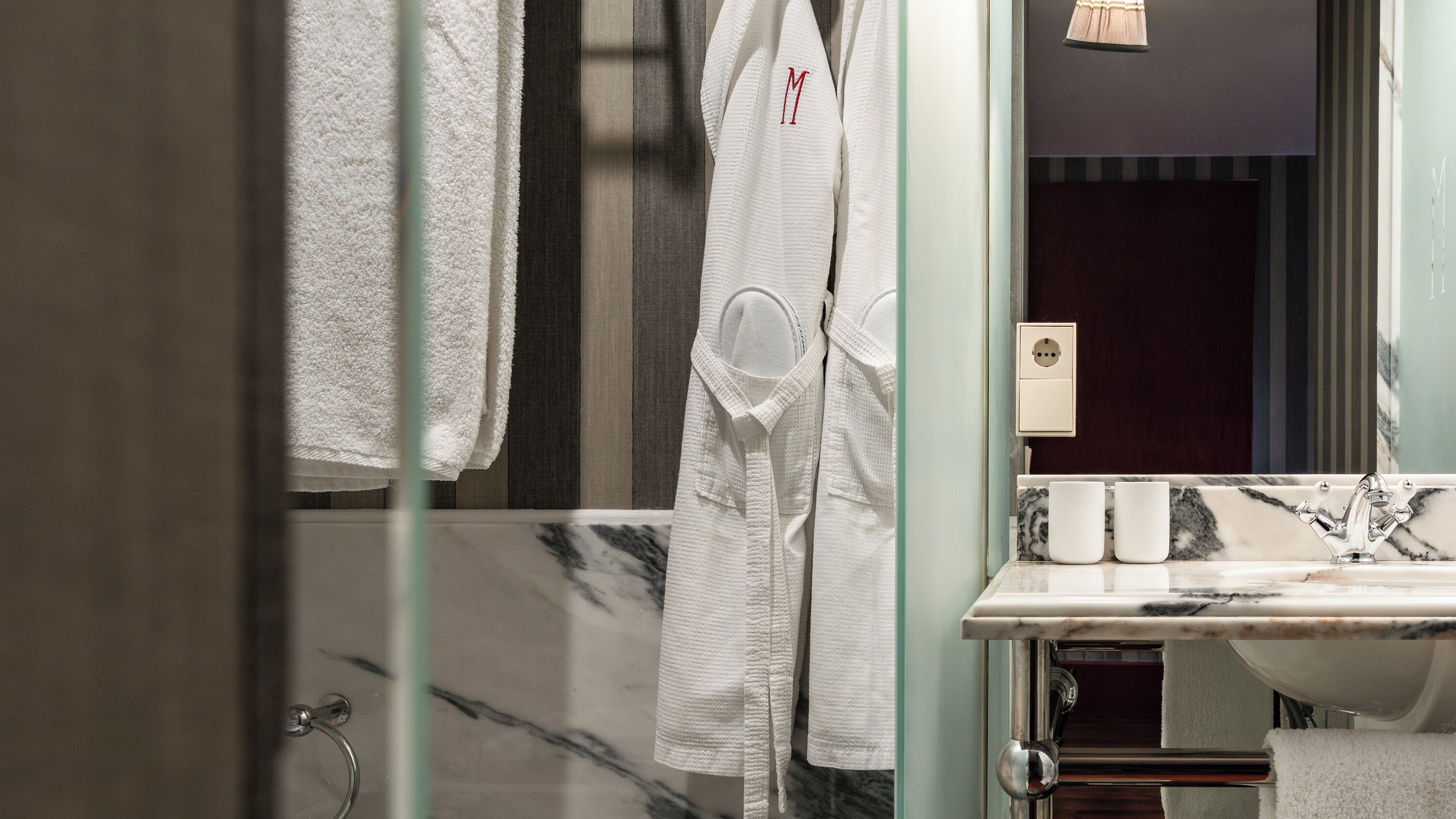 A bathroom with a marble sink, a glass shower enclosure, and a white robe with a red 'M' hanging on the wall.