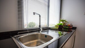A modern kitchen with a stainless steel sink, black countertops, and a window with blinds. - Des Res Hotel and Residence (Samut Prakan)