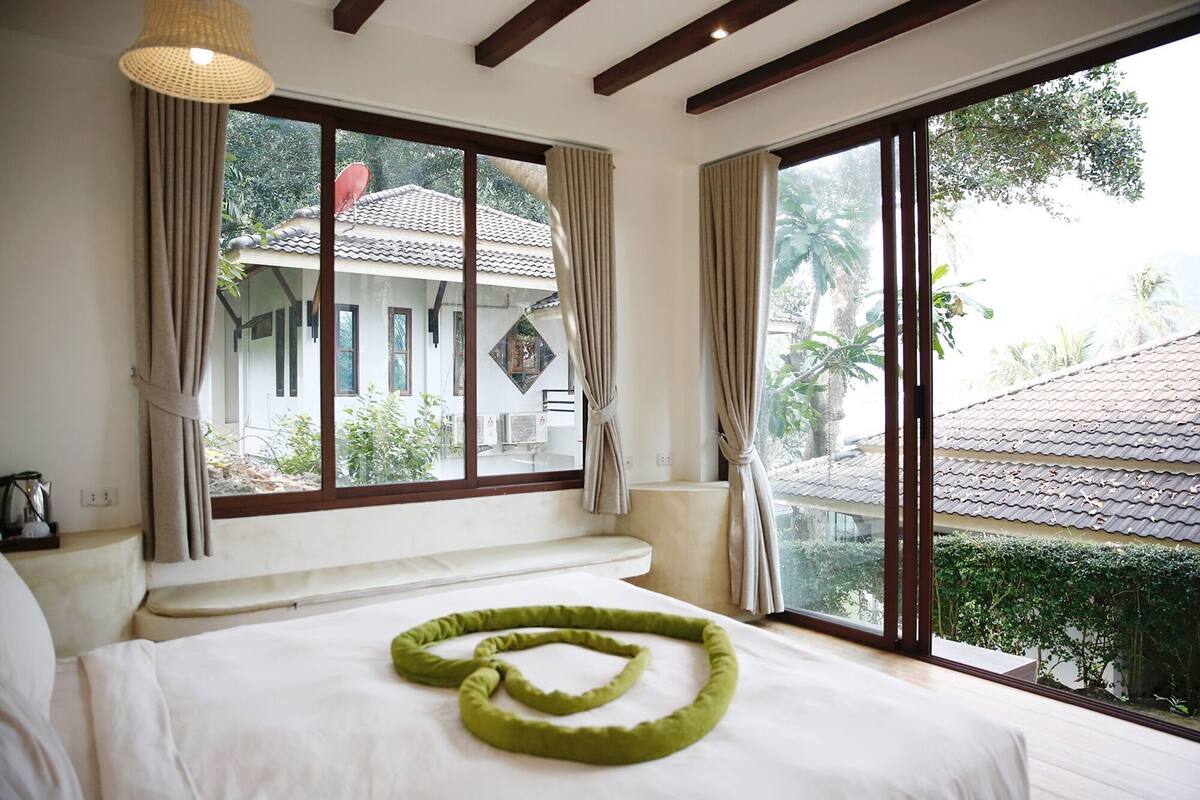 Una habitación con cama, ventanas amplias y vista a un edificio con tejado de tejas.