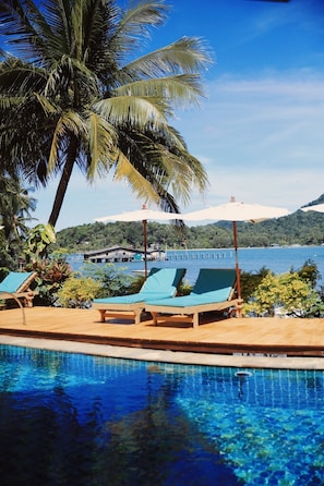 A poolside area with lounge chairs and a view of a body of water and a forested hill in the background.