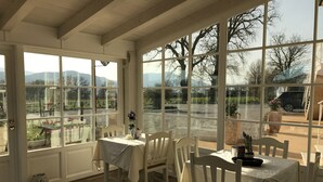 A sunlit dining area with a table set for two, large windows offering a view of the outdoors, and a wooden ceiling.