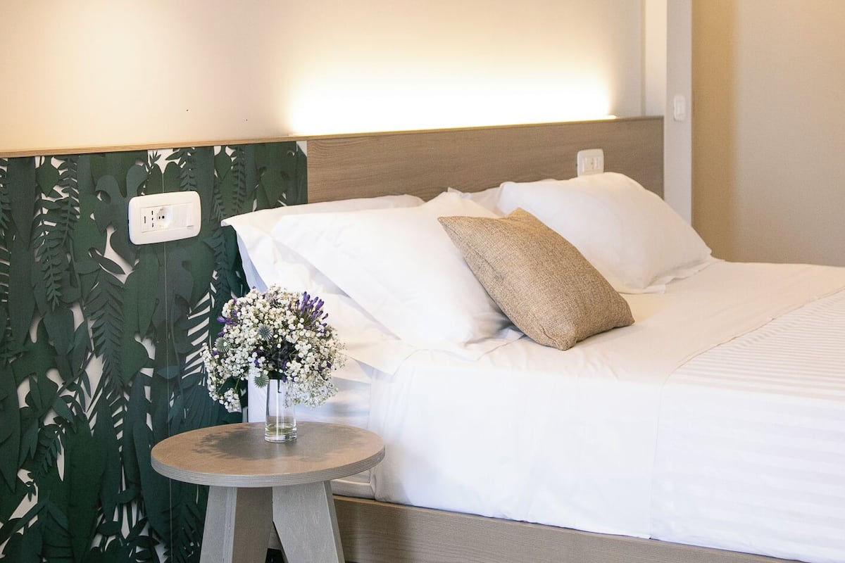 A neatly made bed with white linens and a single decorative pillow, a small round table with a vase of flowers, and a modern pendant light.