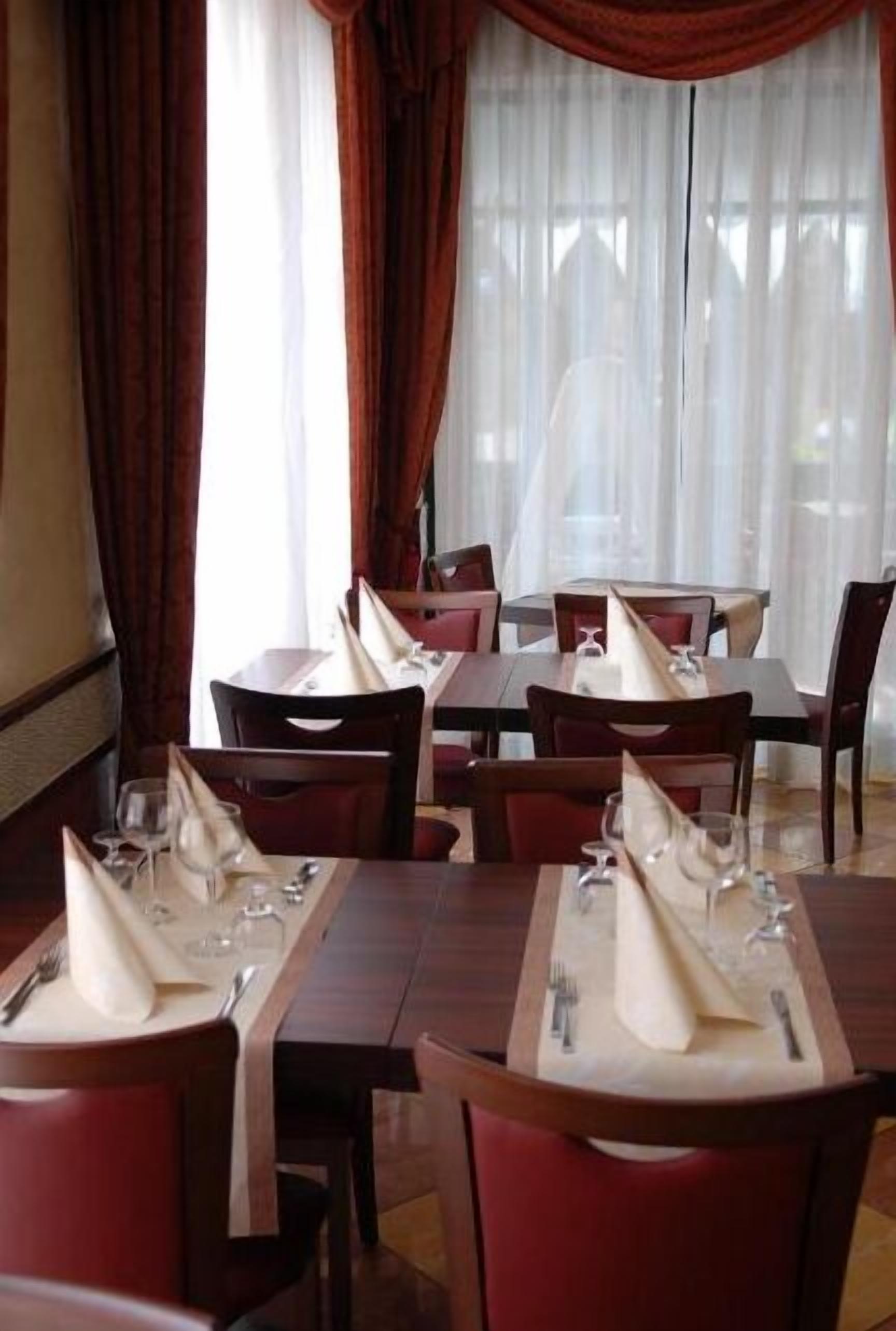 En restaurang med bord dukade med vita servetter, röda stolar och utsikt genom tunna gardiner.
