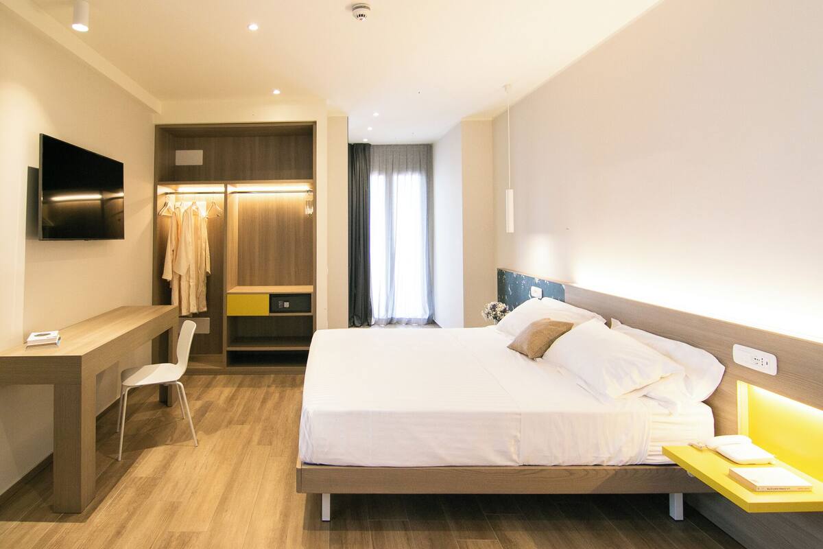 A hotel room with a bed, bedside table, chair, desk, TV, wardrobe, and a window with curtains.