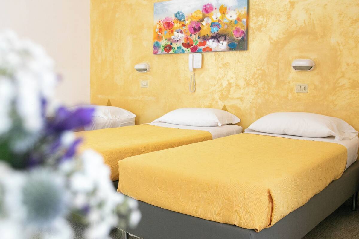 A hotel room with two single beds, each with a yellow bedspread and white pillows. There is a painting of flowers on the wall above the beds.