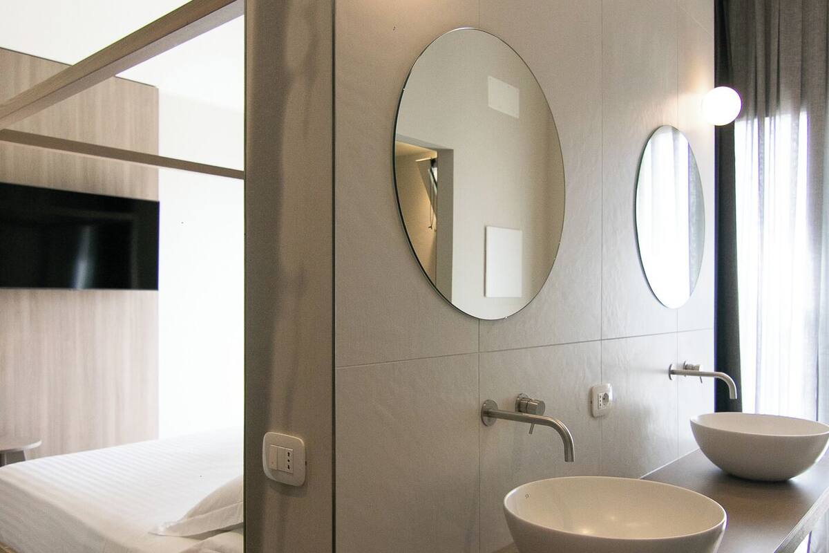 A modern hotel room with a bunk bed, a wooden vanity with a large bowl sink, and two round mirrors.