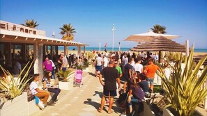 A crowded outdoor area with palm trees, a building with a sign, and a beach in the background. - Hotel Hollywood (Rimini)