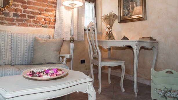 A cozy room with a brick wall, a white sofa, a small table with a tray of flowers, a white chair, and a table with a vase of dried plants. - Casa Del Grande Vecchio (Airasca)