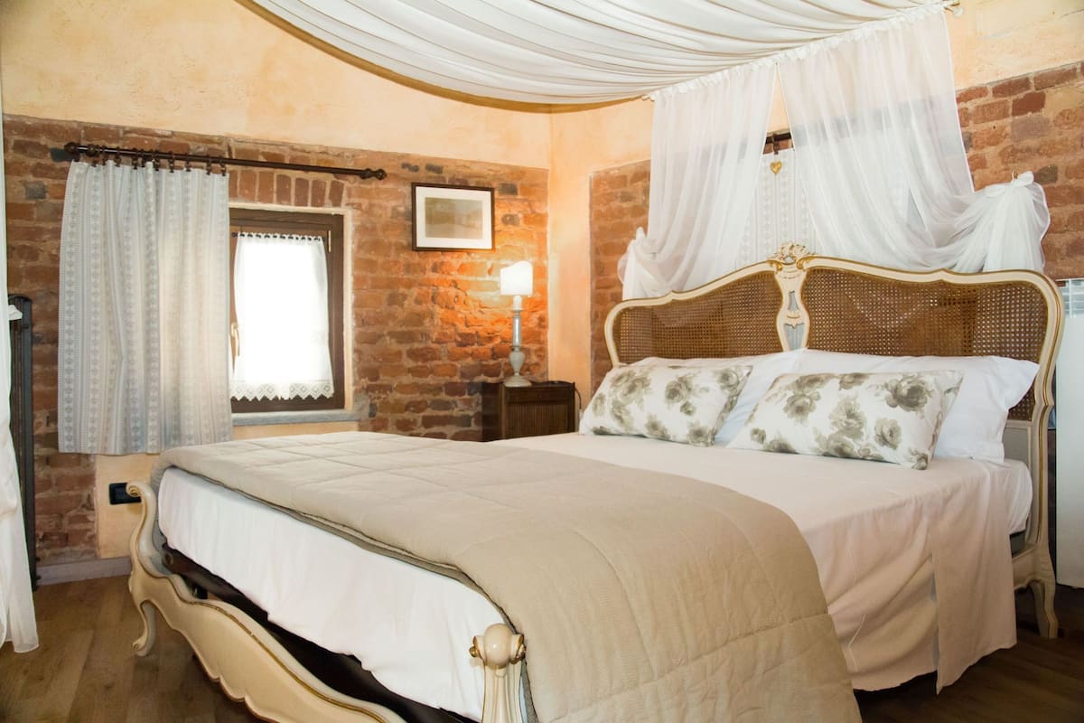 A bedroom with a large bed, a wooden headboard, a nightstand, a wall-mounted lamp, and a window with curtains.