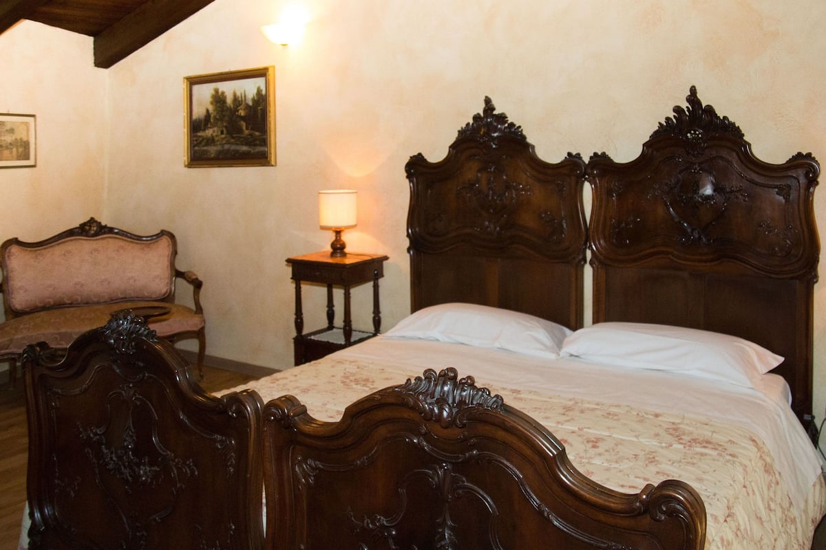 A bedroom with a large wooden bed, a wooden nightstand with a lamp, a wooden chair, and a framed picture on the wall.