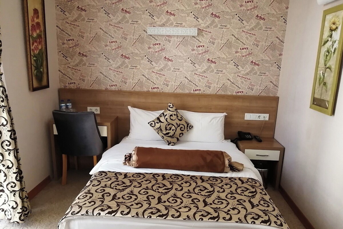 A hotel room with a bed, a desk, a chair, a nightstand, and a wall with a patterned wallpaper.
