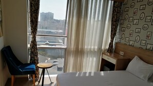 Habitación de hotel con cama, escritorio, silla y vista a un paisaje urbano a través de cortinas translúcidas.