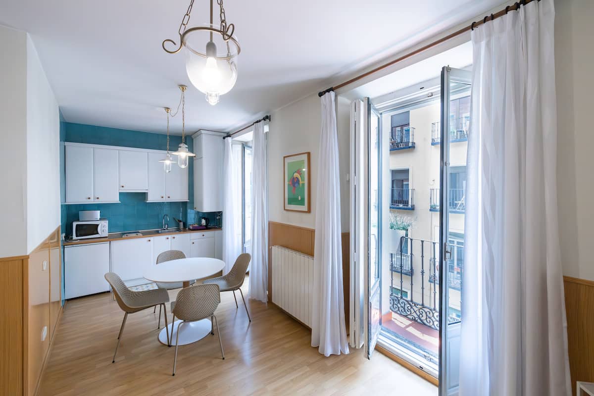 Une cuisine moderne avec des meubles blancs, une table à manger avec des chaises et une vue sur un balcon agrémenté de rideaux blancs.