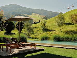 A pool area with lounge chairs and an umbrella, surrounded by lush greenery and a view of a cable car in the distance. - Wiesergut (Saalbach-Hinterglemm)