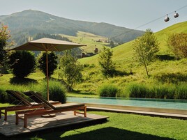 Zona de piscina con sillones de descanso y una sombrilla, rodeada de exuberante vegetación y una vista de un teleférico a lo lejos.