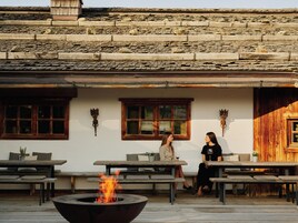 An outdoor seating area with a fire pit, wooden benches, and a building with stone walls and wooden doors.