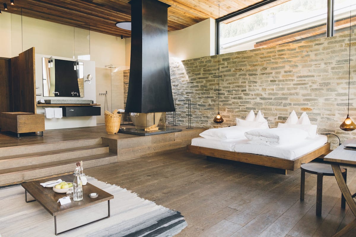 A modern bedroom with a large bed, a wooden ceiling, a stone wall, and a large window.