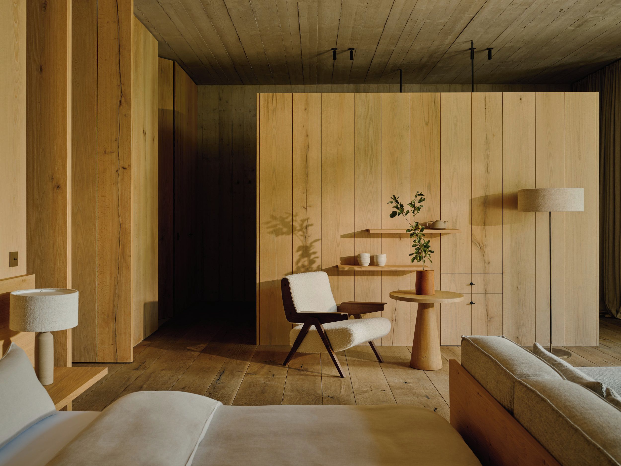 Una habitación moderna con paneles de madera, una cama, una silla, una mesita y una lámpara.