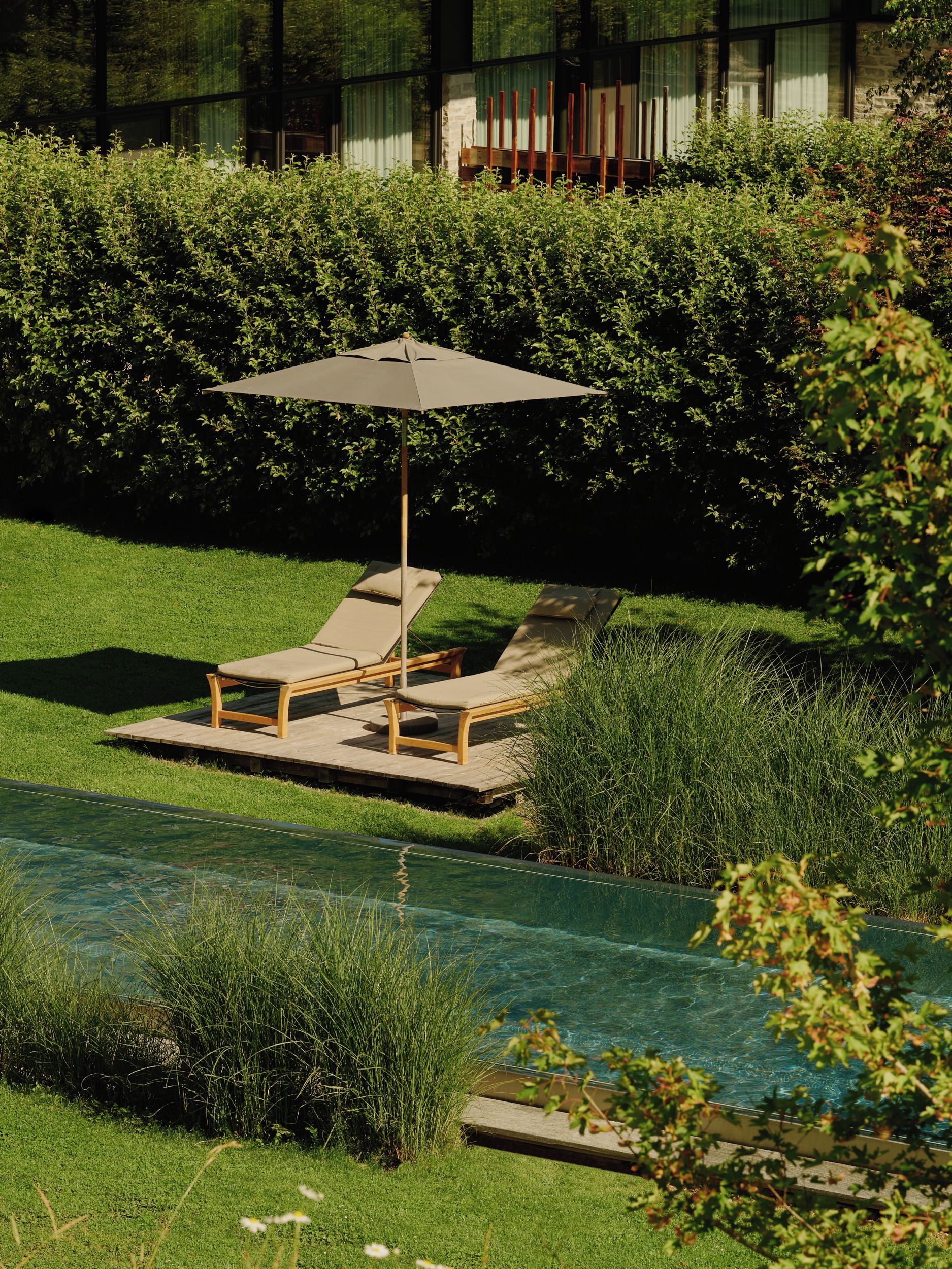 A poolside area with two lounge chairs and an umbrella, surrounded by greenery and a building in the background.