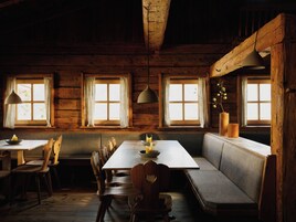 A rustic wooden cabin interior with a dining table, chairs, a bench, and two windows with curtains.