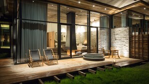 A modern outdoor deck with wooden flooring, glass walls, and a stone wall. There are lounge chairs and a fire pit. - Wiesergut (Saalbach-Hinterglemm)