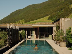A swimming pool with a person in the water, surrounded by a wooden deck and a building with large windows. - Wiesergut (Saalbach-Hinterglemm)