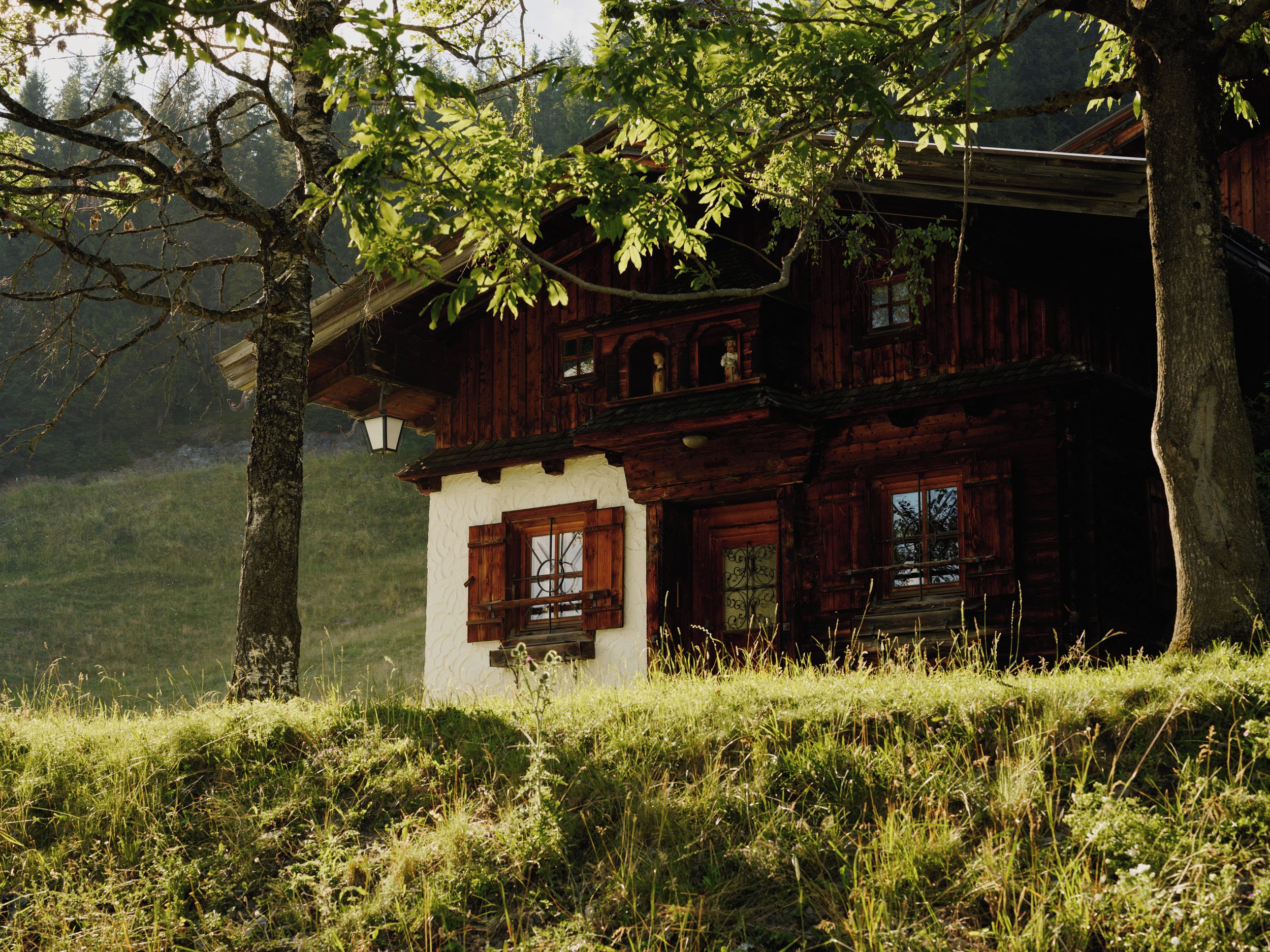 Une maison en bois à l’allure rustique, dotée d’un balcon et entourée d’une végétation luxuriante et d’arbres.