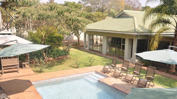 A swimming pool with lounge chairs and umbrellas, surrounded by a garden and a house with a tiled roof. - Coghlan Villa Guest House (Harare)