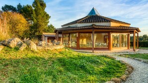 Un belvédère de style traditionnel, doté d’un toit en tuiles et de poutres en bois, entouré d’un chemin de gravier et d’une végétation luxuriante.