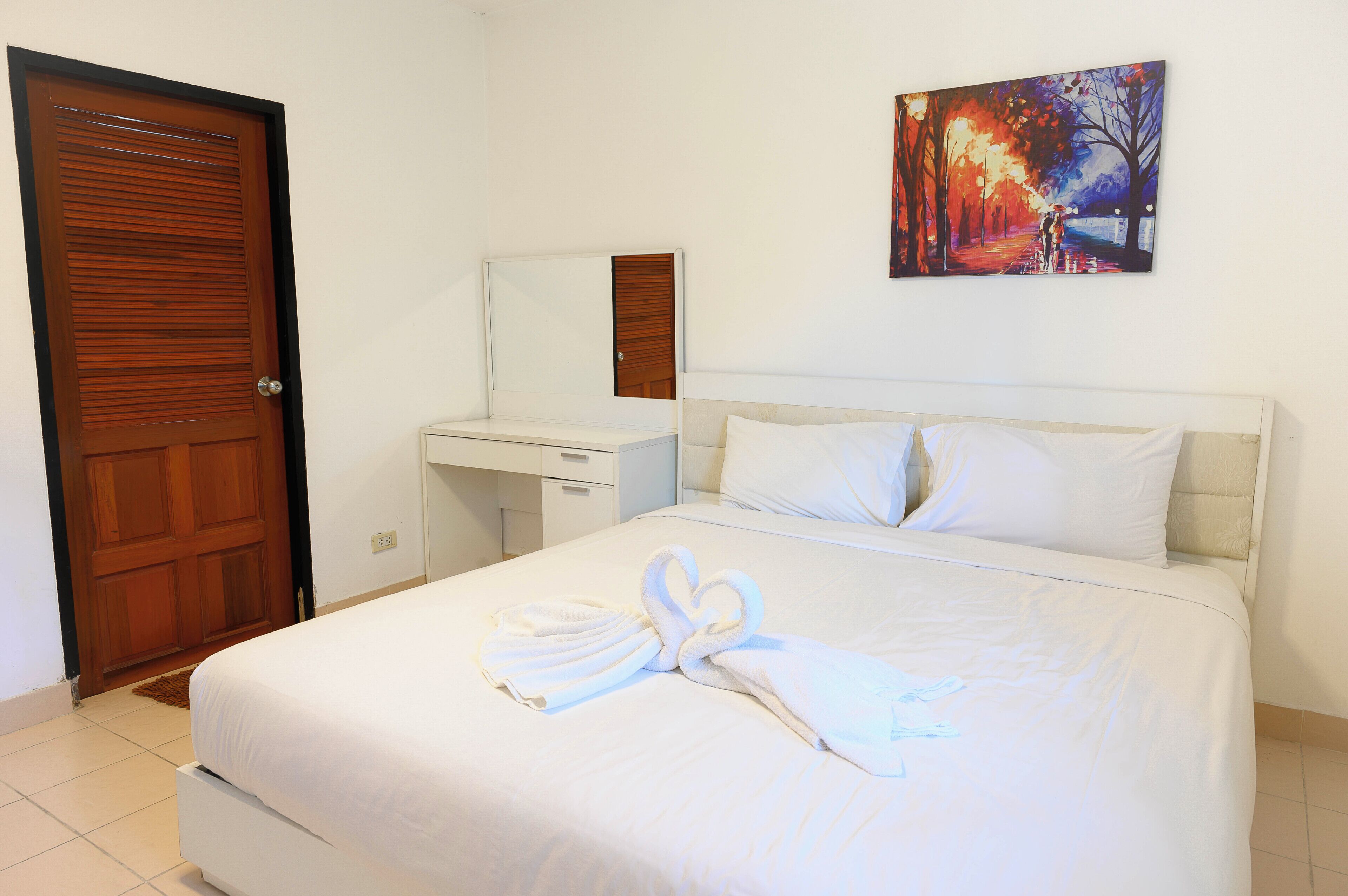 Une chambre d’hôtel avec un lit, une porte en bois, une table de chevet blanche, un miroir et un tableau accroché au mur.