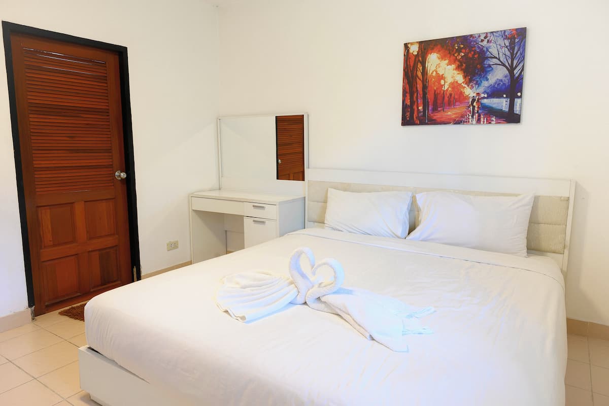 A hotel room with a bed, a wooden door, a white bedside table, a mirror, and a painting on the wall.