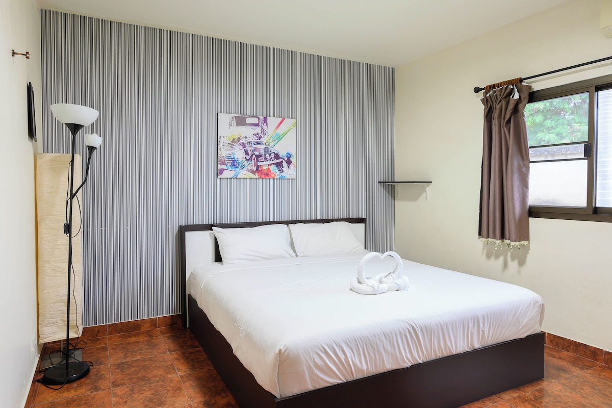 A hotel room with a bed, a wall-mounted artwork, a floor lamp, and a window with curtains.