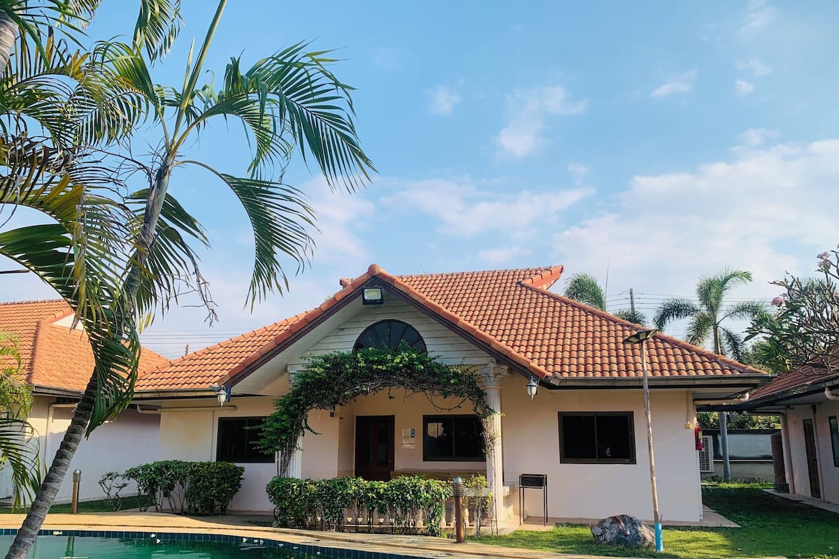 A two-story house with a tiled roof, a swimming pool, and palm trees.