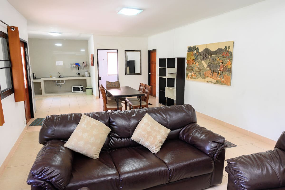 A living room with a brown leather sofa, a dining table with chairs, and a kitchen area in the background.