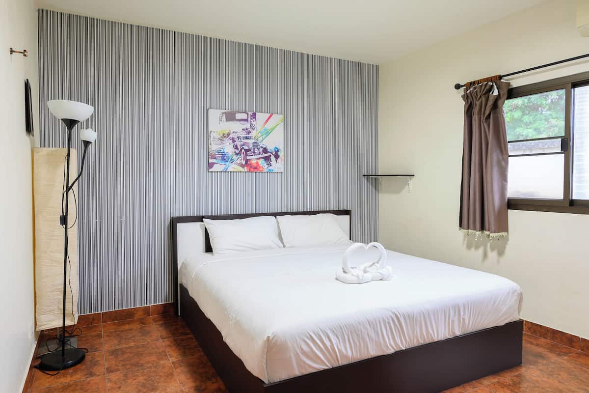 A hotel room with a bed, a wall-mounted artwork, a floor lamp, and a window with curtains.