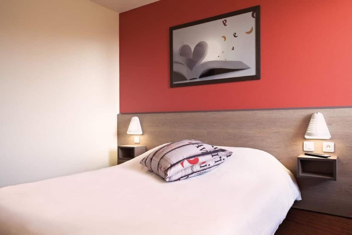 Een hotelkamer met een bed, kussens, een nachtkastje met een lamp en een lijst met een foto aan de muur.